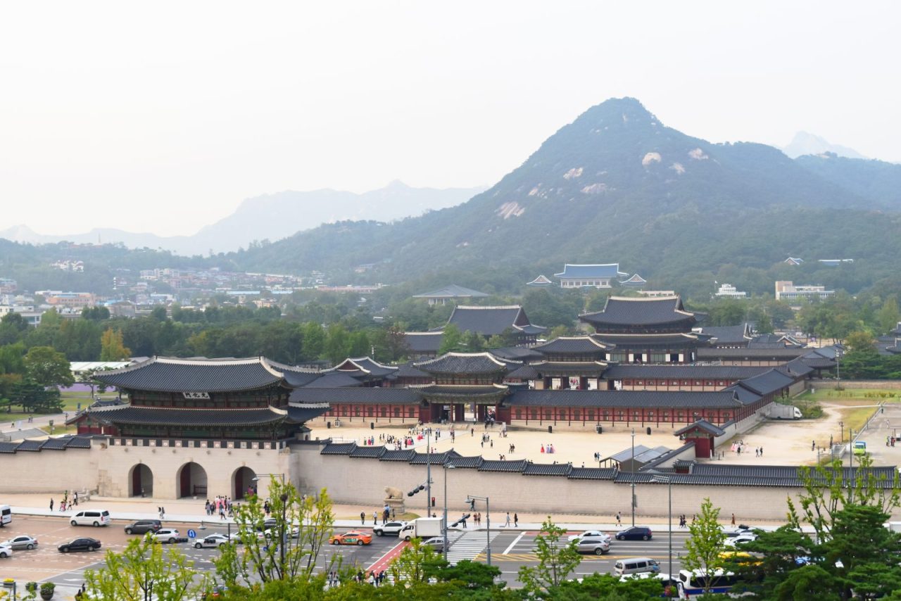 Gyeongbokgung Palace and its tiled gates, with a busy Seoul road in front and mountains behind.