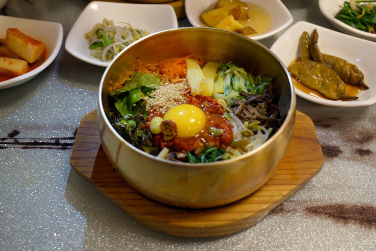 Bibimbap topped with egg yolk, vegetables and sesame seeds, with side dishes behind it.