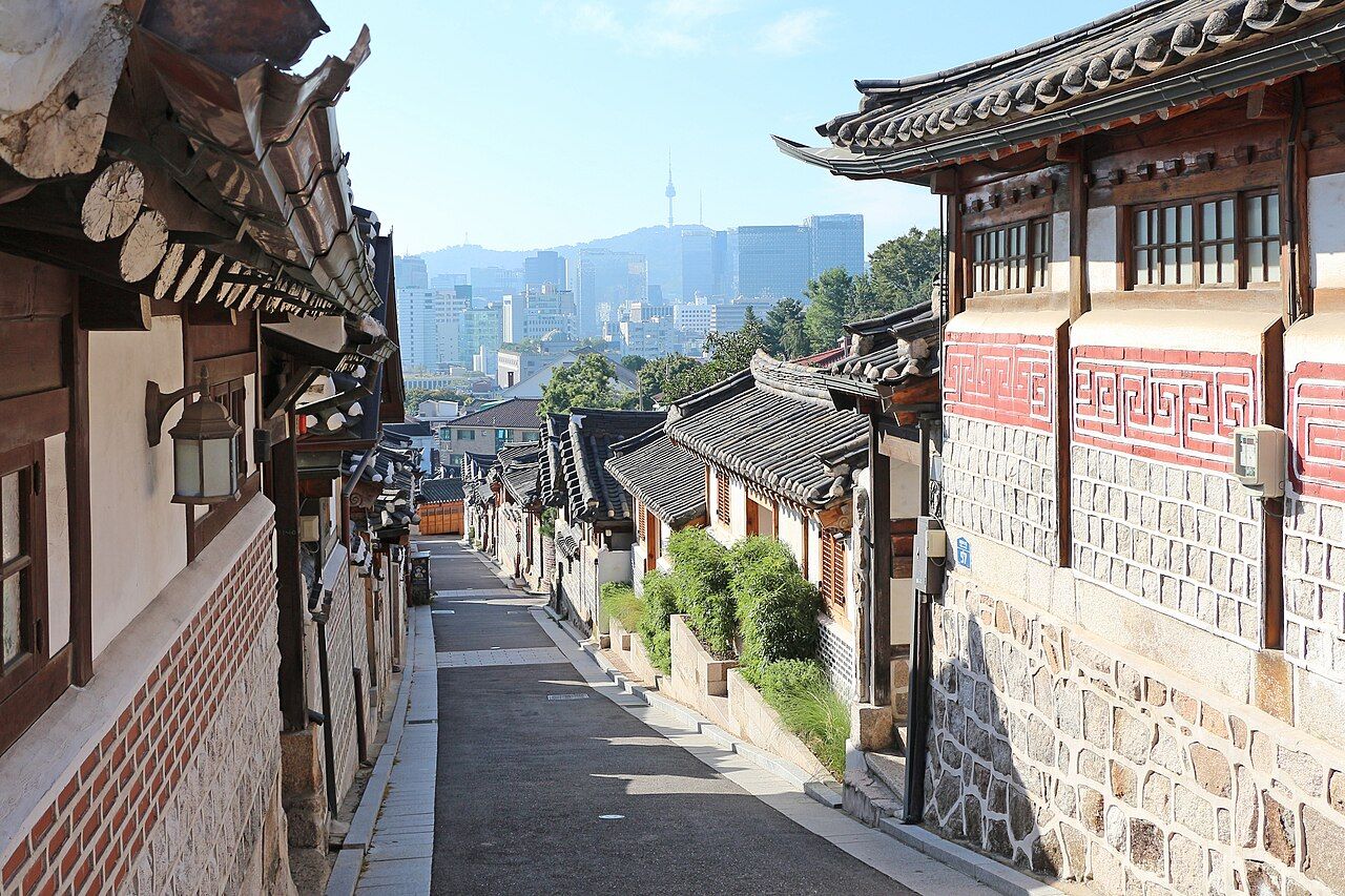 An empty sloping lane between tiled hanok houses in Bukchon, with central Seoul and N Seoul Tower in the distance.